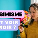 Femme en pull jaune tenant des lunettes de soleil noires devant un mur turquoise, pour illustrer le pessimisme et voir tout en noir