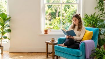 Solitude choisie : une femme lit seule et sereine dans un fauteuil turquoise près d'une fenêtre